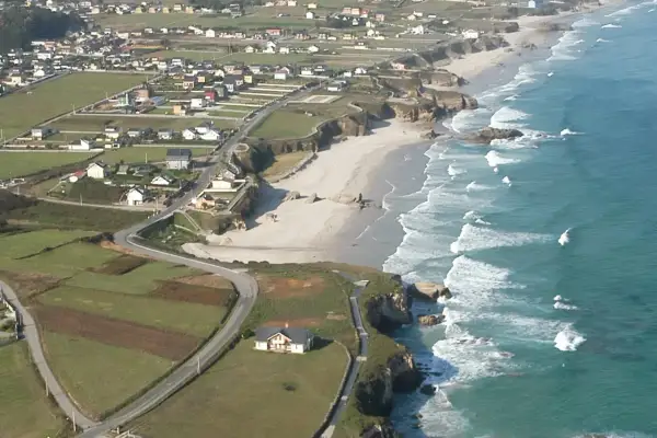 Vista aérea de la playa Longara en Lugo, con casas blancas y campos verdes bordeando la costa.