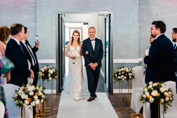 Panna młoda w sukni ślubnej idzie z ojcem do ołtarza, trzymając bukiet.