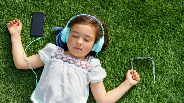 Dziewczynka w słuchawkach leży na trawie, słuchając ulubionej piosenki na Dzień Dziecka. Obok telefon i okulary.