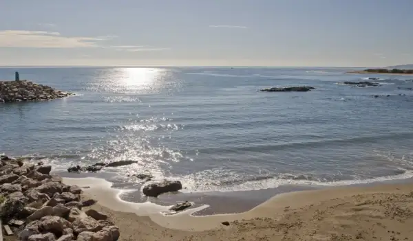 Costa de Villaricos con un faro en un rompeolas de rocas. El sol brilla sobre el mar azul y la arena dorada.
