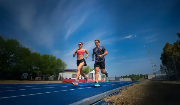 Coppia corre su pista blu, un allenamento intenso per la loro prossima corsa a tappe. Cielo azzurro e nuvole bianche.