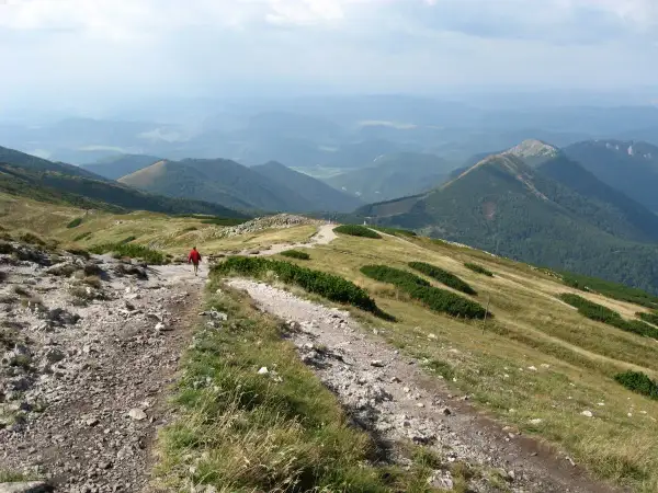 Wędrówka po górach Słowacji na weekend. Widok na zielone wzgórza i doliny, kamienista ścieżka, samotny turysta w czerwonej kurtce.