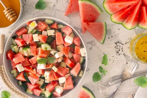 Erfrischender Wassermelonen Feta Salat mit Gurkenstücken und Minze, garniert mit Pfeffer. Daneben ein Glas Dressing und Honig.