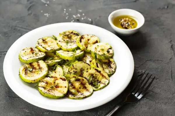 Zucchine alla scapece non fritte, grigliate e condite con olio e spezie, servite su un piatto bianco con una ciotolina di condimento.
