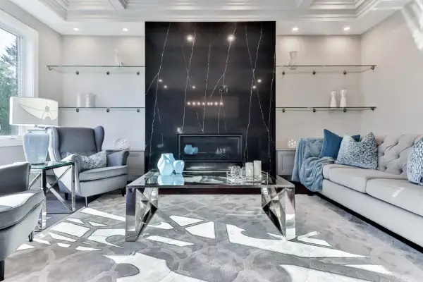 Elegancki salon z kominkiem obłożonym czarnym marmurem i **ścianami w stylu glamour**. Szare fotele, sofa i szklane półki tworzą luksusową atmosferę.
