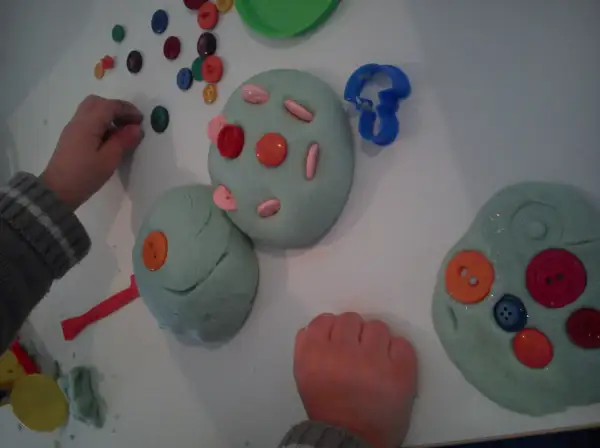 Manos infantiles jugando con plastilina casera, decorada con botones de colores y cortadores.