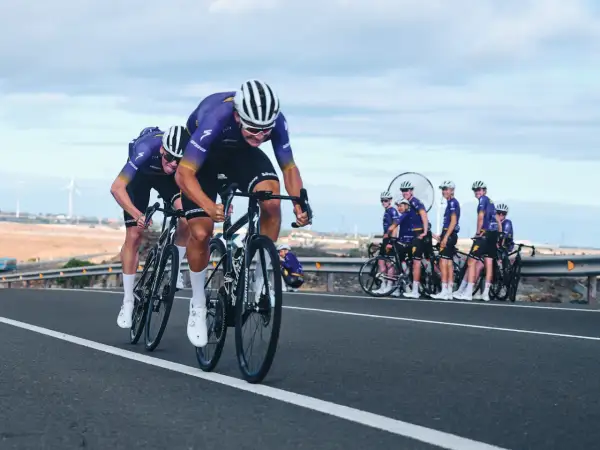 Cykla på Gran Canaria i tävlingsform. Ett cykellag i lila dräkter susar fram på en asfalterad väg med vindkraftverk i bakgrunden.