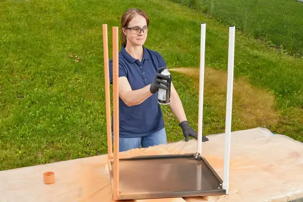 Kvinna sprayar metallbenen på ett bord utomhus. Hon bär skyddsglasögon och handskar.