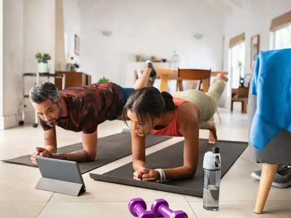 Ein Paar macht Liegestütze zu Hause, inspiriert von **micro workouts** auf einem Tablet. Hanteln und Wasserflasche liegen bereit.