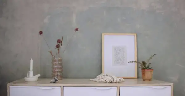 Un tavolino con candela, vaso di fiori secchi, quadro e corda. Sembra la preparazione calce per imbiancare, con un tocco artistico.