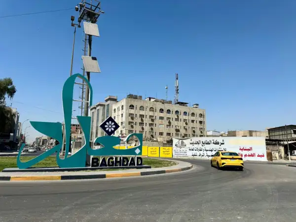 Wielki napis "BAGHDAD" i stylizowany napis arabski na tle budynków i nieba. Stolica Iraku wita podróżnych.