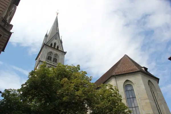 Der Turm der Stephanskirche Konstanz ragt mit seiner Spitze in den blauen Himmel. Ein Baum mit dichtem Laub steht davor.