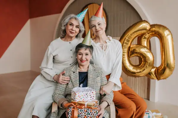 Eleganckie stylizacje na urodziny w lokalu. Trzy kobiety świętują 60. urodziny, otoczone prezentami i złotymi balonami.