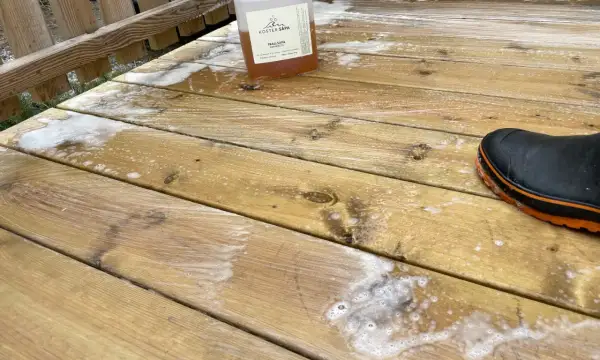 En flaska linoljesåpa står på en altan som håller på att rengöras. En svart stövel med orange kant syns i nedre högra hörnet.