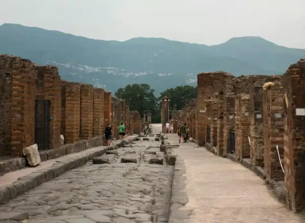 Besucher spazieren auf der antiken, kopfsteingepflasterten Straße von Pompeji heute, umgeben von den Ruinen römischer Gebäude. Berge im Hintergrund.