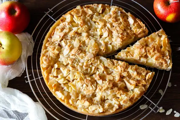 Ein goldbrauner florentiner apfelkuchen mit knusprigen Mandelblättchen, daneben Äpfel und ein Stück Kuchen.