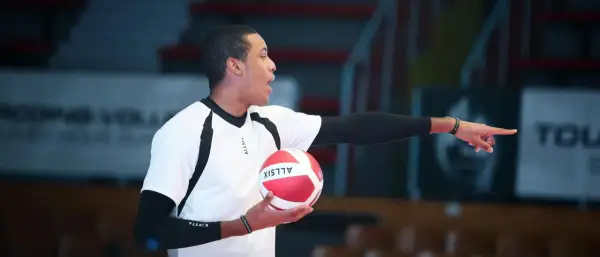 Młody siatkarz z piłką w dłoni, wskazujący palcem, jakby tłumaczył zasady gry, która narodziła się z potrzeby aktywnego spędzania czasu.