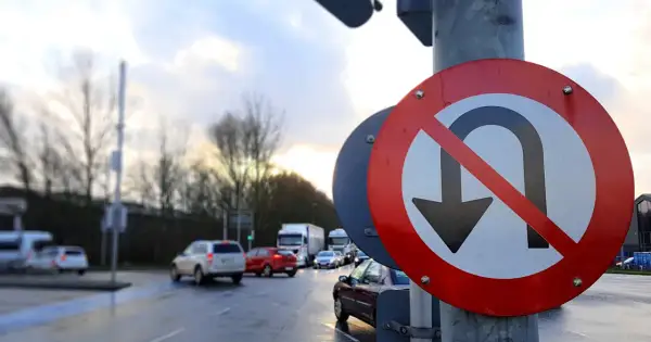 Znak zakaz zawracania na mokrej drodze, z samochodami w tle. Kierowcy muszą uważać, bo nie wolno tu zawracać.