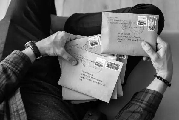 Dłonie trzymają stos brązowych kopert z adresami, znaczkami i pieczątkami pocztowymi. Jedna z kopert jest zaadresowana na "Paige Newell", sugerując odbiór poste restante.