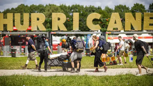 Besucher mit Rucksäcken und Gepäckwagen auf dem Hurricane Zeltplatz. Große goldene Buchstaben "HURRICANE" im Hintergrund.
