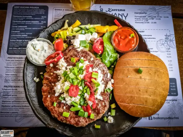 Soczysty kotlet z dodatkami i bułką, serwowany z sosem. Prawdziwa uczta kuchni bałkańskiej.