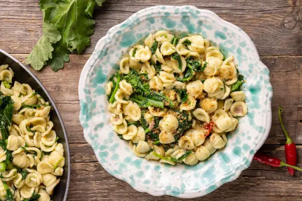 Orecchiette con cime di rapa, la ricetta originale pugliese, servite in un piatto decorato, con peperoncini rossi e foglie di rapa sullo sfondo.