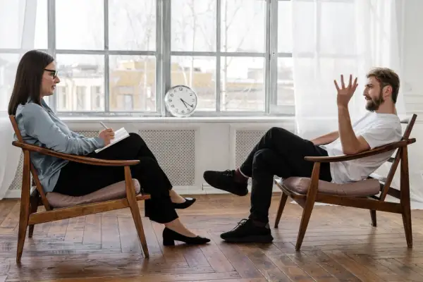 Uomo e donna seduti su sedie di legno, in conversazione. La donna scrive su un quaderno. La prima seduta psicologo si paga, un investimento per il benessere.