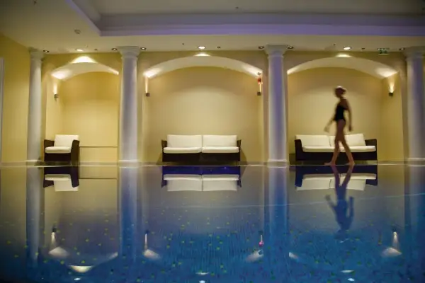 Una mujer camina junto a una piscina interior, rodeada de sofás y columnas, creando una atmósfera de relajación para una experiencia spa sensorial.