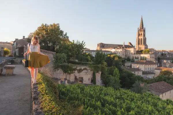 Donna passeggia su un muro a Saint-Émilion, con il villaggio e la sua chiesa in lontananza.