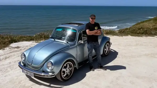 Man står bredvid en ljusblå Volkswagen Bubbla vid havet. En påminnelse om vem grundade Volkswagen och dess ikoniska design.
