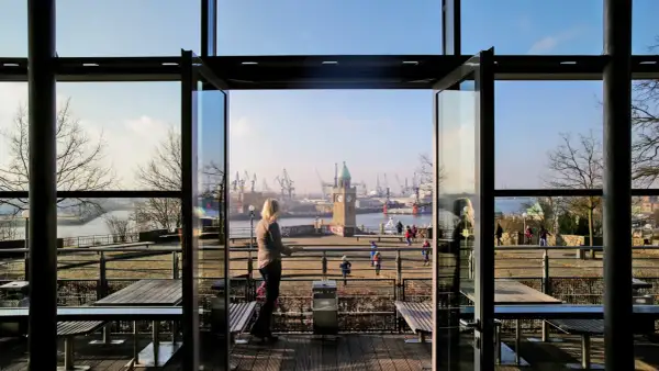 Blick durch offene Glastüren auf den Hamburger Hafen mit dem alten Elbtor und Kränen. Eine Frau genießt die Aussicht, perfekt für einen Stintfang Hamburg.