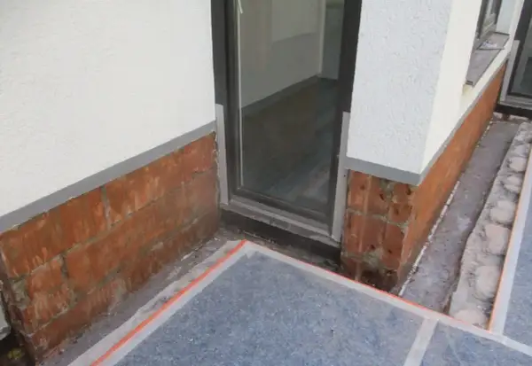 Rote Ziegelsteine am Sockel zeigen Anzeichen von aufsteigender Feuchtigkeit im Sockelbereich. Ein Fensterrahmen ist sichtbar.
