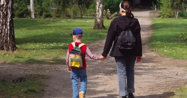 Dzieci autystyczne lubią spacery i poczucie bezpieczeństwa. Chłopiec z plecakiem idzie za rękę z kobietą po ścieżce w parku.
