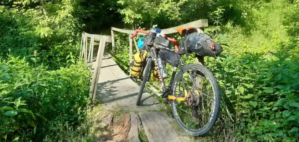 Rower wyprawowy na drewnianym moście, gotowy na mazowiecki gravel. Otoczony bujną zielenią, zaprasza do przygody.