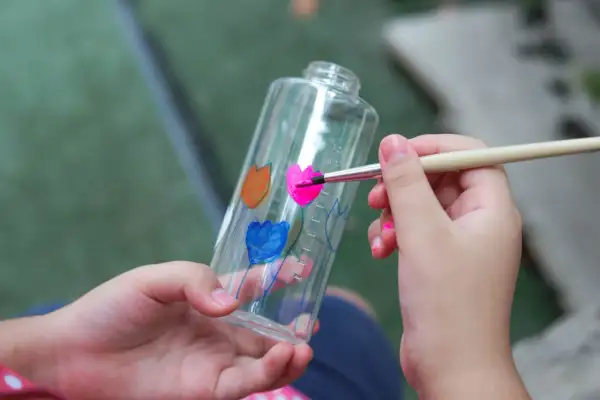 Bambino dipinge fiori su una bottiglia trasparente, un modo creativo per imparare come colorare la plastica trasparente.