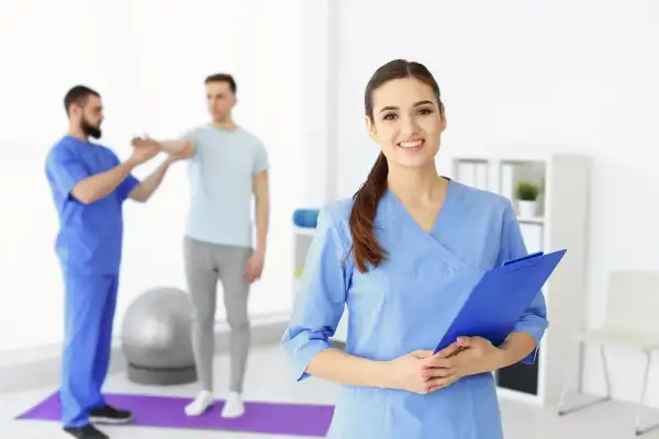 Fizjoterapeutka w niebieskim uniformie trzymająca teczkę, w tle terapeuta ćwiczący z pacjentem.