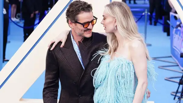Pedro Pascal y su pareja, con ella luciendo un vestido azul con flecos, se miran tiernamente en una alfombra azul.