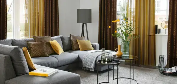 Gemütliches Wohnzimmer mit grauem Sofa, Kissen in Braun und Gelb, sowie moderne Fenstergestaltung mit zweifarbigen Vorhängen.