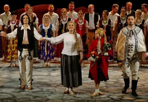 Grupa ludzi w strojach ludowych, mężczyzna w białej koszuli, kamizelce i spodniach z haftem, kobieta w kwiecistej sukni i halka.