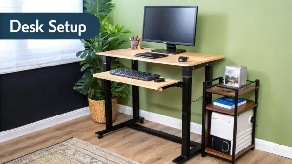 Adjustable height computer desk setup with monitor, keyboard, and mouse. A small side table holds a computer tower and books.
