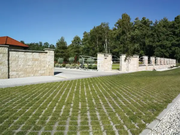 Piękny, kamienny mur i brama wjazdowa, a przed nimi zielona przestrzeń z ażurami na drogę.