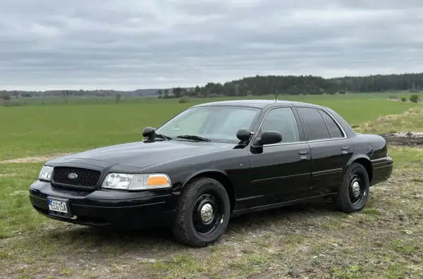 Svart Ford Crown Victoria, en robust bil som står parkerad på en grusväg med gröna fält och skog i bakgrunden.