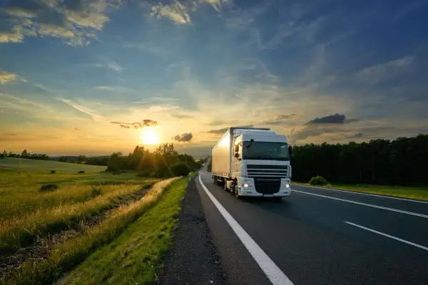Un camion blanc roule sur une route au coucher du soleil, traversant une campagne verdoyante. C'est le quotidien de ceux qui travaillent sur la route.