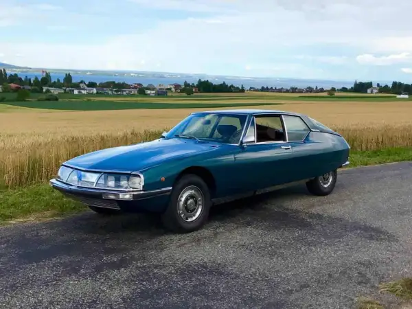 Piękny, turkusowy Citroen SM na tle złotego pola i jeziora. Klasyk motoryzacji w malowniczej scenerii.