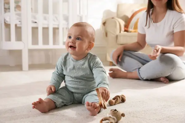Uśmiechnięte 10 miesięczne dziecko bawi się drewnianymi zabawkami na dywanie, podczas gdy mama siedzi w tle.