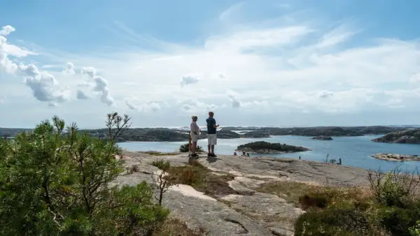 Två personer njuter av utsikten från Vetteberget i Fjällbacka. Vackert väder med moln och sol över skärgården.