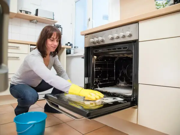 Frau mit gelben Handschuhen reinigt den Backofen. Schaum und ein blauer Eimer stehen bereit.