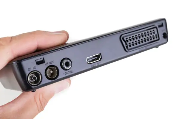 Dłoń trzyma czarny dekoder z portami RF IN, RF OUT, COAXIAL, HDMI OUT i SCART.