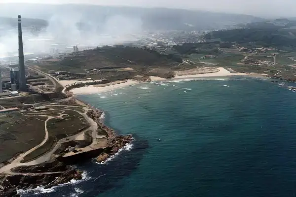 Vista aérea de la playa de Sabón, con una chimenea industrial y humo en el fondo, y el mar azul turquesa.