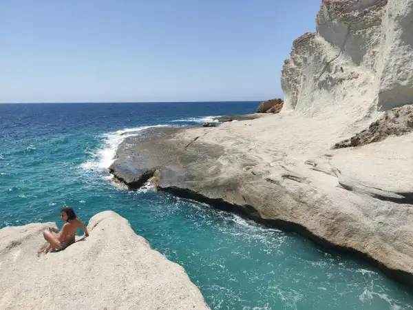 Una chica sentada en una roca en una cala chica, con el mar azul turquesa y acantilados blancos al fondo.